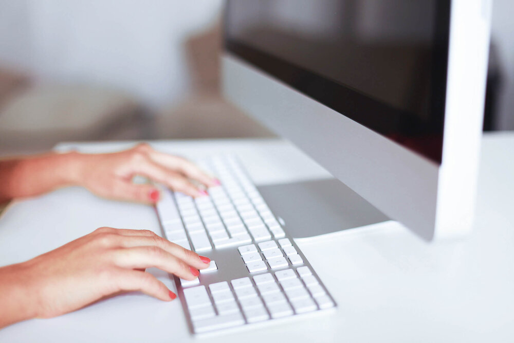 Zwei Frauenhände tippen auf einer Tastatur, die vor einem iMac-Computer steht.