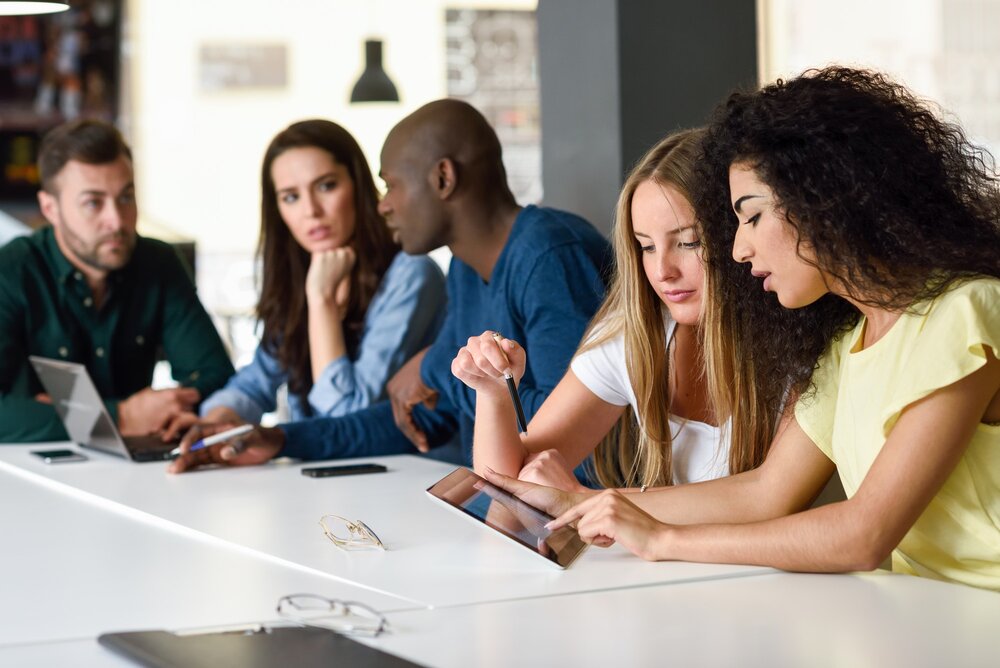 Mehrere junge Menschen sitzen nebeneinander an einem Tisch und diskutieren mit Hilfe eines Tablets und eines Laptops über ein Schulungsthema.